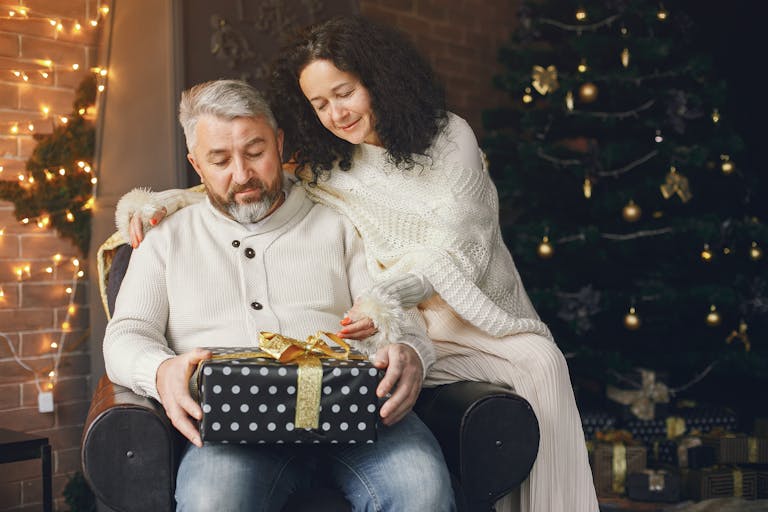 A happy couple in warm sweaters enjoying a Christmas present by the fireplace and tree.