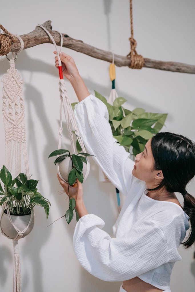 A woman arranges hanging plants in macrame holders against an indoor wall.