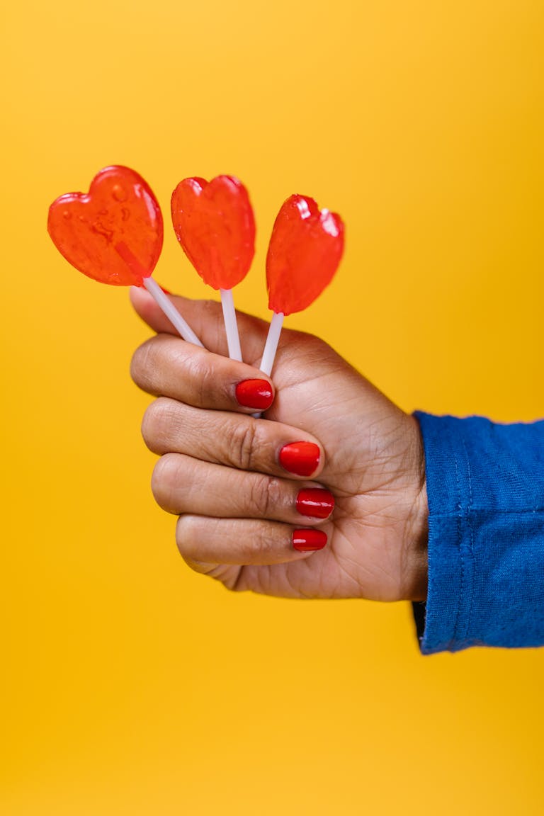 A hand with red nails holding three heart-shaped lollipops on a vibrant yellow background, perfect for Valentine themes.