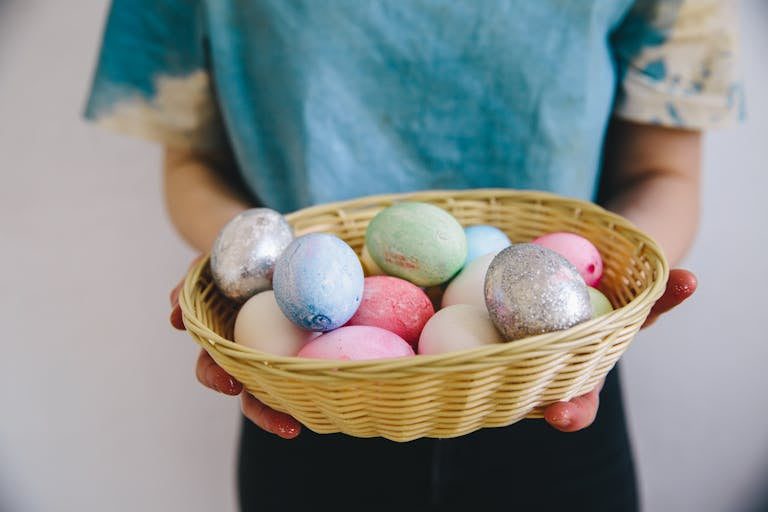 A person holding a basket filled with vibrant Easter eggs in pastel and metallic colors.