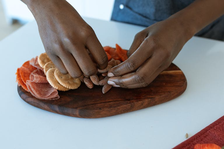 Close-up of hands arranging a charcuterie board with almonds, meats, and crackers on a wooden tray.