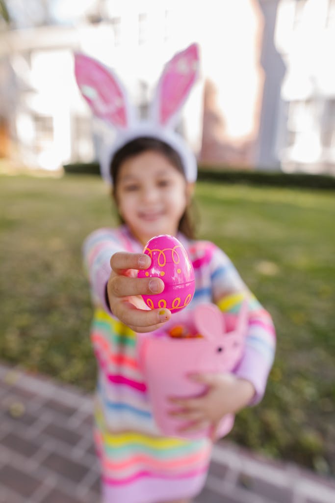 Smiling girl with bunny ears holding a decorated Easter egg outdoors.