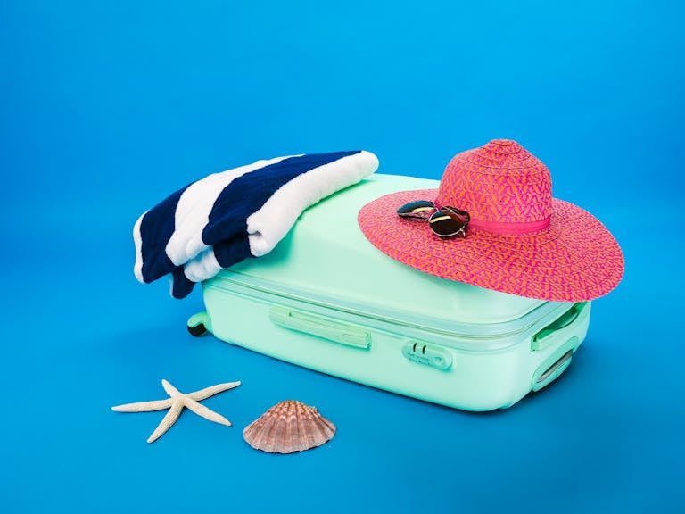 Beach essentials including a suitcase, hat, towel, and seashells on a bright background.