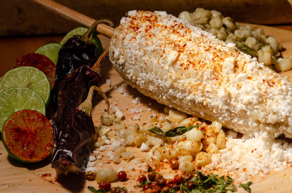 Delicious Mexican elote served with limes and dried chili peppers on a wooden board.