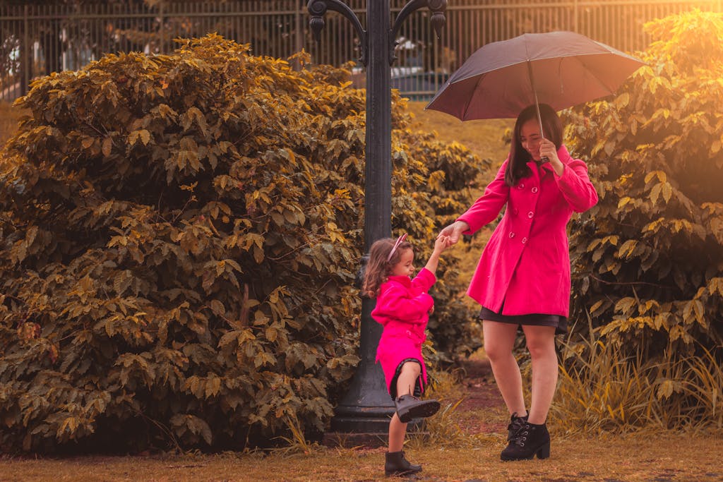 A mother and daughter enjoy a sunny outdoor walk under an umbrella in matching pink coats.