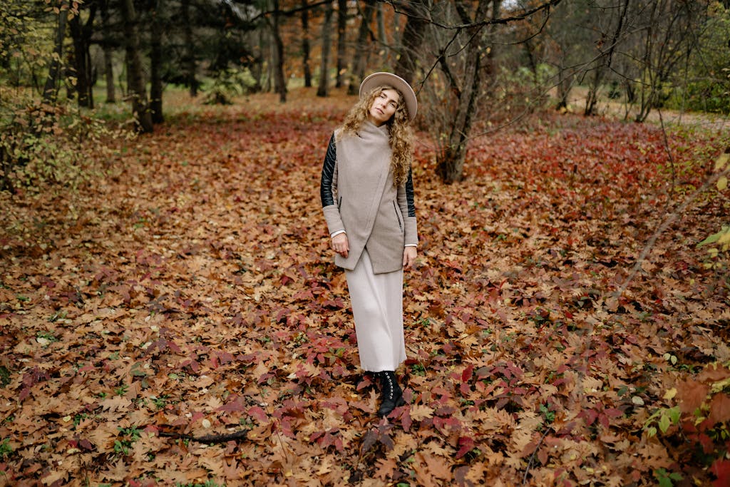 Stylish woman in warm clothing poses amidst colorful autumn leaves in a serene forest setting.