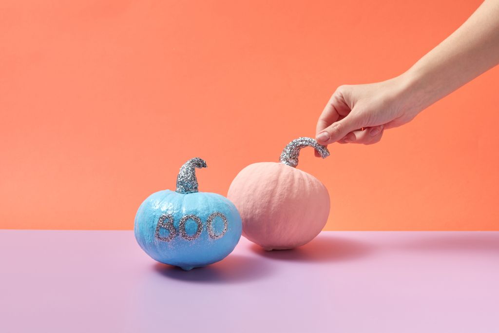 Hand holding pink pumpkin with glitter stem next to blue pumpkin with 'boo' text, orange background.